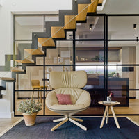 Beige armchair with a pink accent cushion on a dark blue rug, set against modern glass and wood staircase.