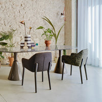 Modern dining table with a clear glass surface, paired with gray and patterned green chairs against a natural stone wall.