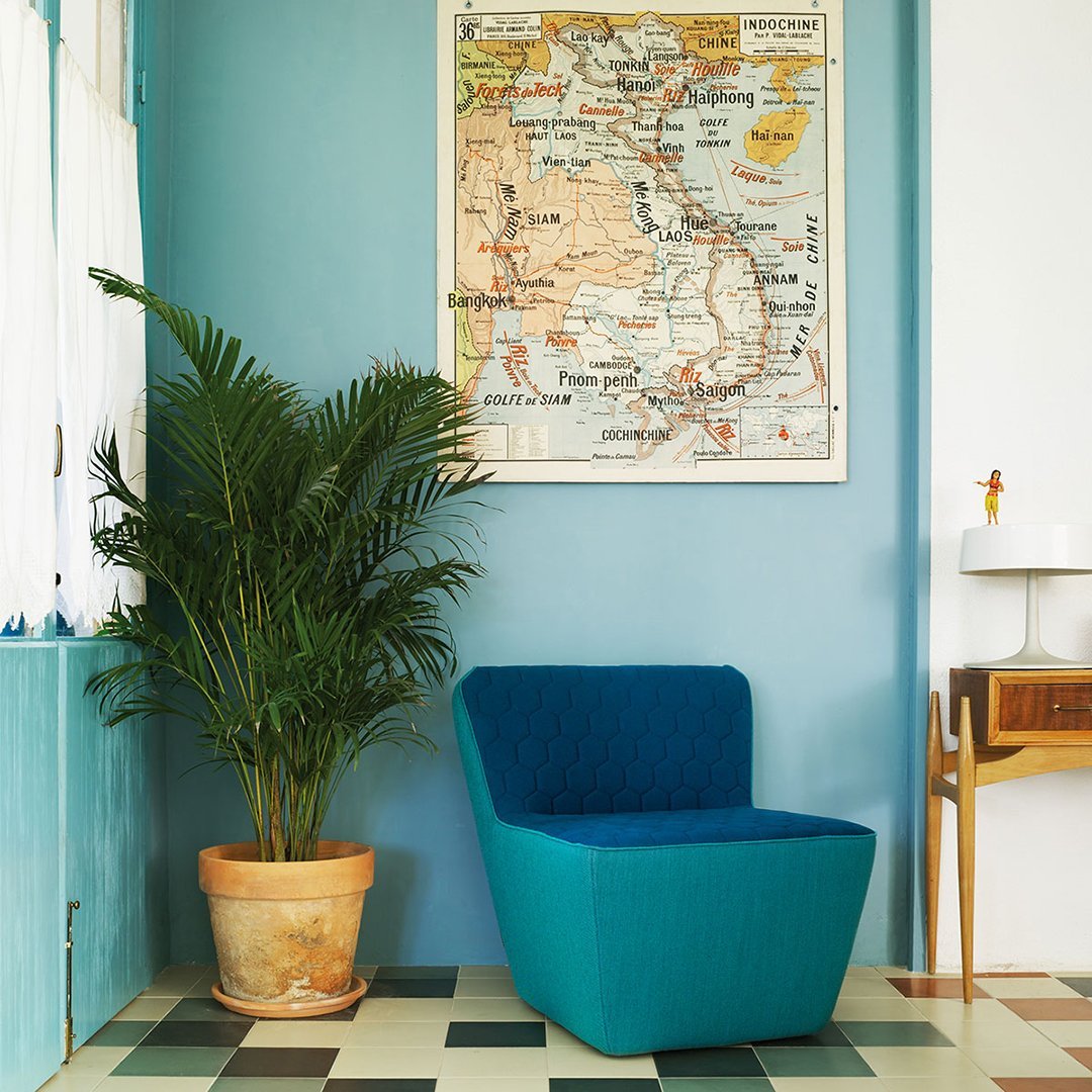 Teal upholstered chair against light blue wall, green plant, and patterned checkered floor tiles in muted tones.