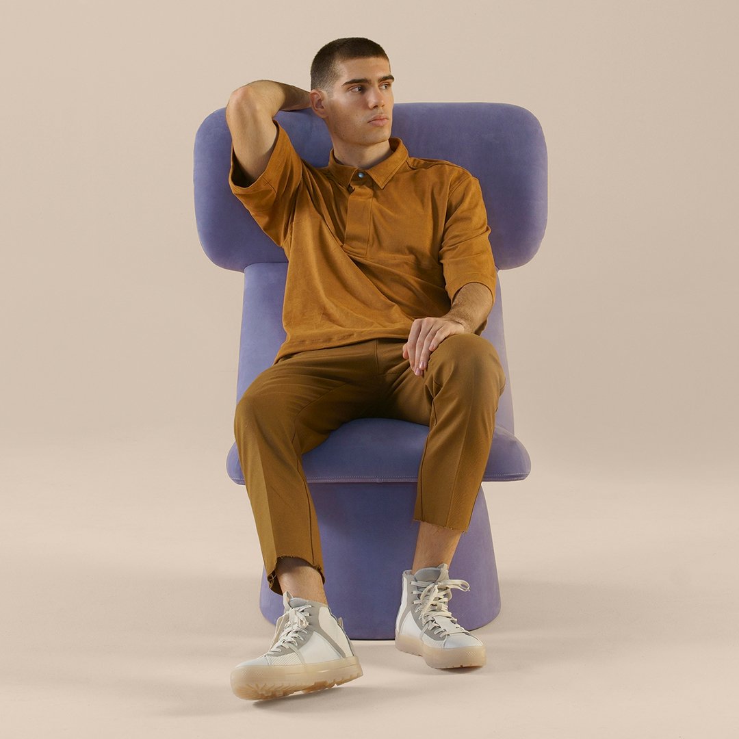 Man seated in a purple chair, wearing a mustard-colored polo and brown pants, paired with white sneakers.