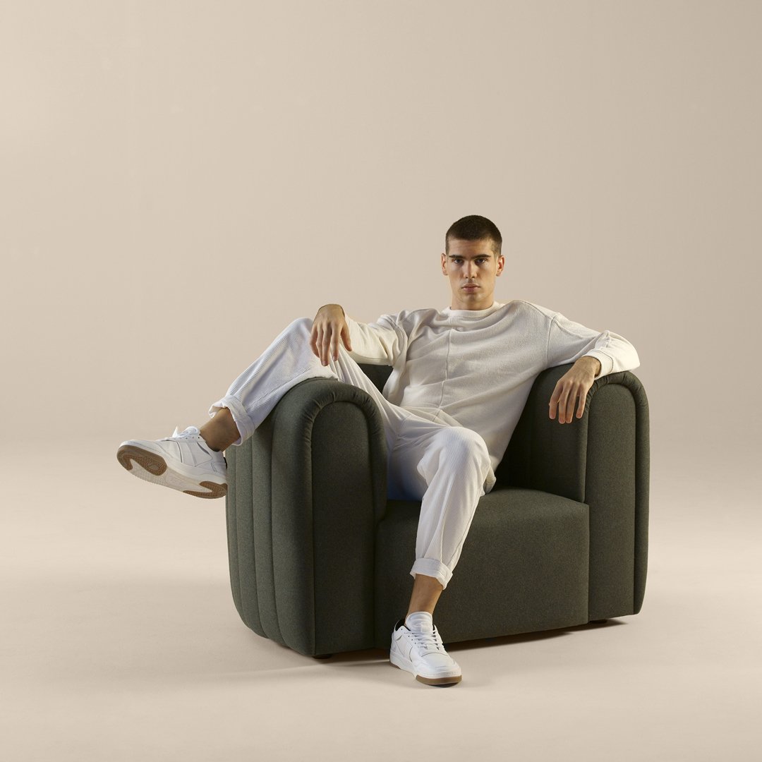 Stylish green upholstered chair with smooth curves, paired with white clothing and sneakers against a neutral backdrop.