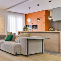 Modern living room featuring beige sofa with green and patterned pillows, orange accent kitchen, and sleek wooden elements.