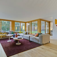 Modern living room featuring a rich burgundy rug, soft gray sofas, colorful cushions, and bright wooden accents.