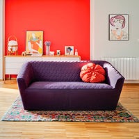 Vibrant purple sofa with geometric texture, accented by a bold orange pillow, on a colorful patterned rug against a red wall.