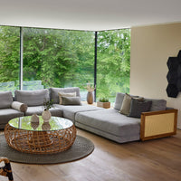 Cozy gray sectional sofa with textured pillows, woven rattan coffee table, and natural light-filled green backdrop.