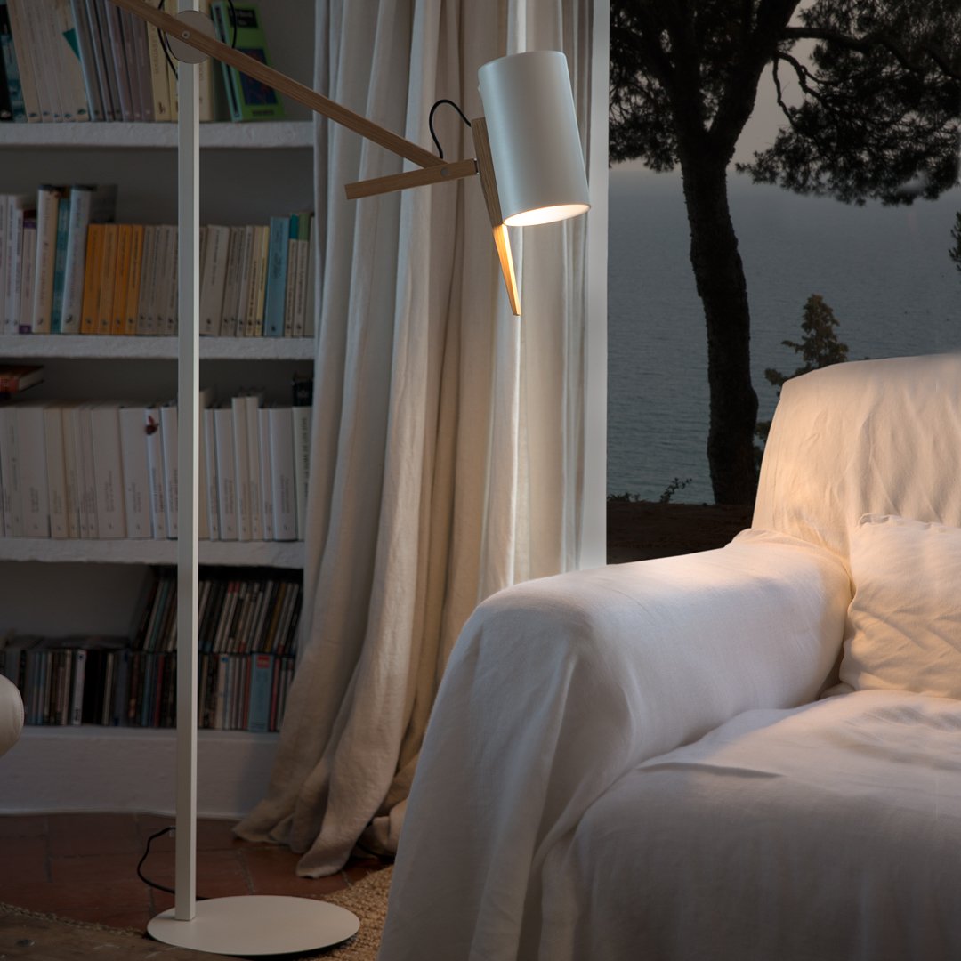 Modern white floor lamp with wooden accents, positioned near a light-colored couch in a cozy, softly-lit setting.