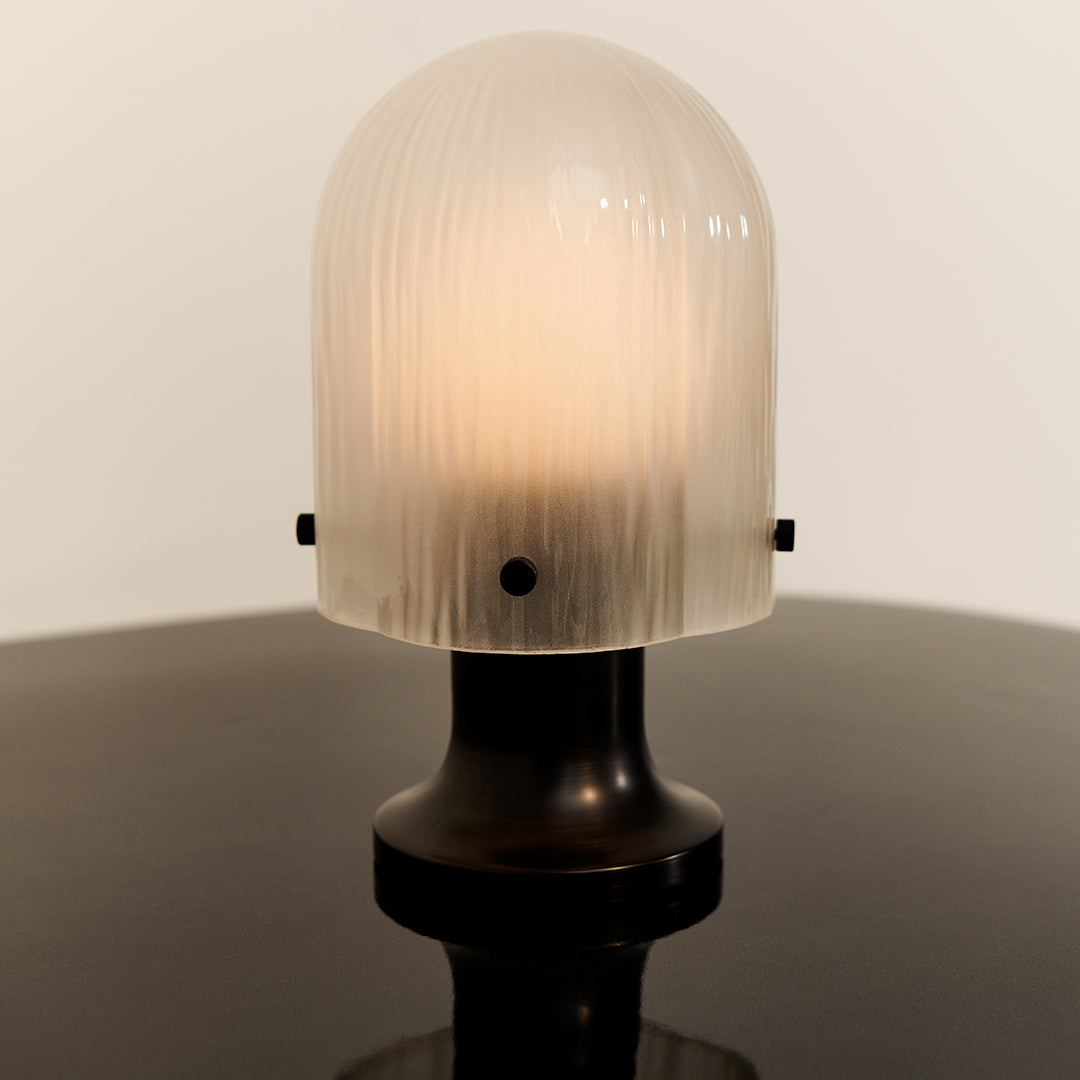 "Frosted glass lamp with ribbed texture, black base, glowing warm light, minimalist design on a glossy surface."
