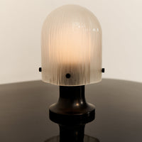 "Frosted glass lamp with ribbed texture, black base, glowing warm light, minimalist design on a glossy surface."
