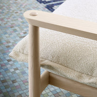 Light beige cushion with a textured pattern, resting on a natural wood chair, against a colorful tiled floor backdrop.
