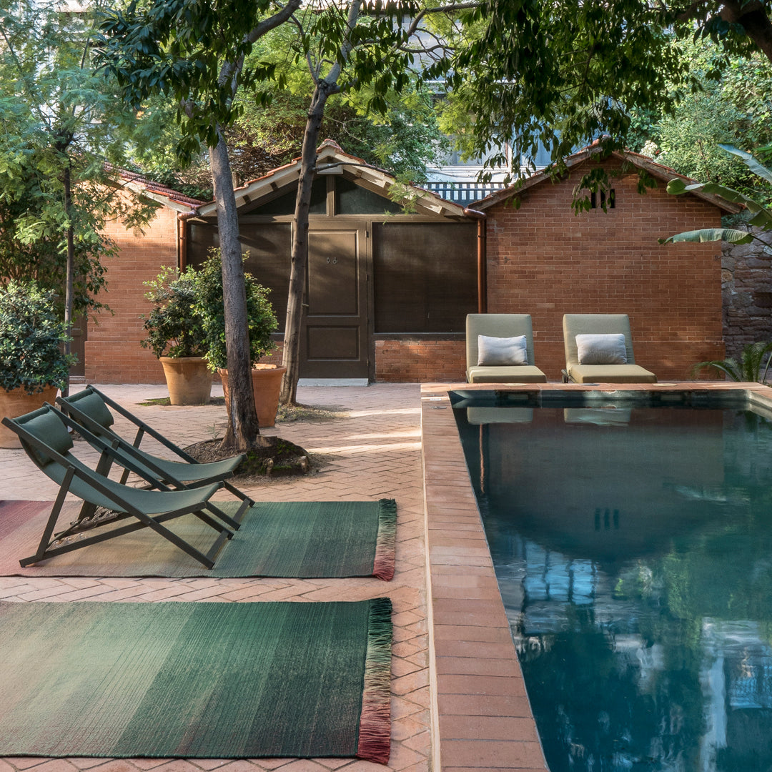 "Stylish green and burgundy outdoor rugs beside a tranquil pool, surrounded by lush greenery and warm brick tones."