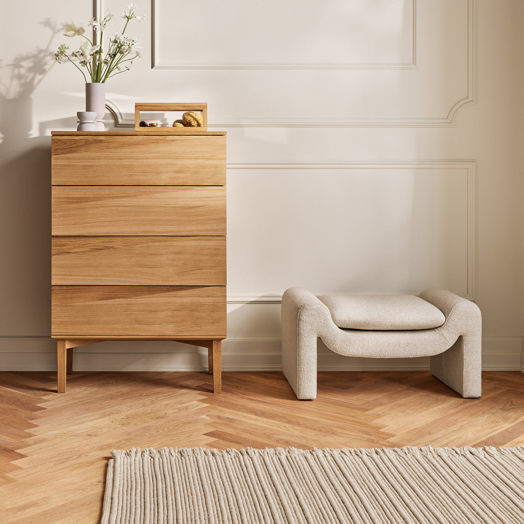 Stylish wooden dresser in natural oak with beige upholstered stool and striped cream rug on herringbone floor.