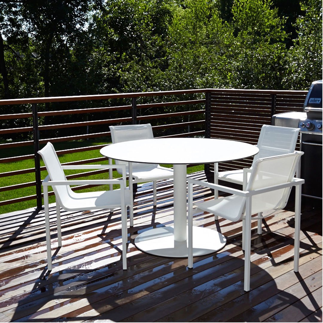 White circular table with four matching chairs on a wooden deck surrounded by lush greenery.