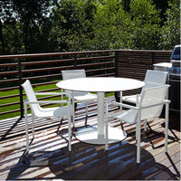 White circular table with four matching chairs on a wooden deck surrounded by lush greenery.