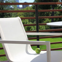 White patio chair with a smooth fabric design, set against a lush green background and sleek wooden railing.