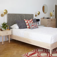 "Light wood bed with gray textured headboard, vibrant red and blue patterned pillows, and a geometric red and cream rug."