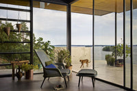 Modern lounge chair in green with a blue accent pillow, surrounded by greenery and natural wood tones, bright and airy setting.