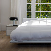 Sleek white bedding on minimalist bed frame with a light wood floor and clear windows, creating a serene, modern look.