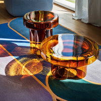 Amber glass side tables with smooth curves, set on a vibrant abstract patterned rug featuring blue, purple, and orange tones.
