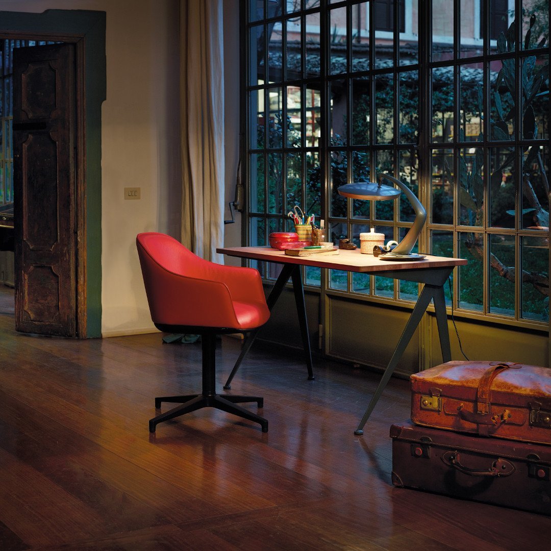 Red chair with smooth curves, wooden desk with a natural finish, large window framing greenery, warm wood floors.