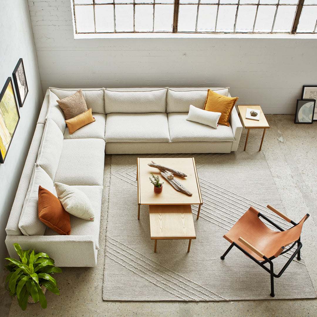 Light beige sectional sofa with orange and brown accent pillows, beige rug, and wooden tables in a modern setting.