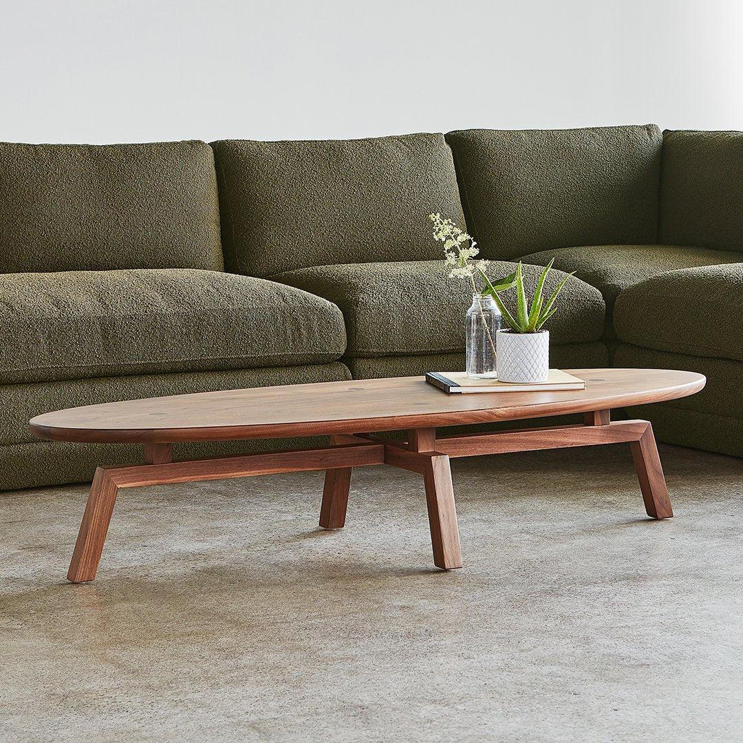 Oval wooden coffee table in warm brown, paired with an olive green sofa, set on a light gray floor. Simple and modern design.