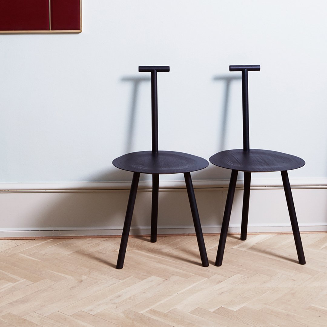 Black minimalist chairs with round seats and unique handle-back design, set against a light wood floor and pale wall.