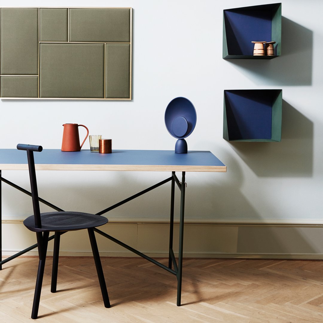 Modern workspace featuring blue tabletop, black chair, olive green wall art, and geometric navy shelves.