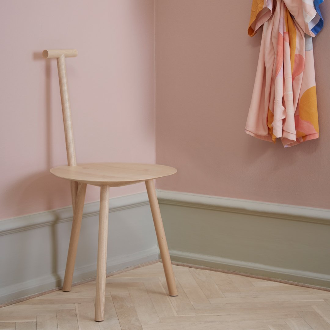 Light wood chair with a minimalist design, set against soft pink walls and patterned fabric drapes.