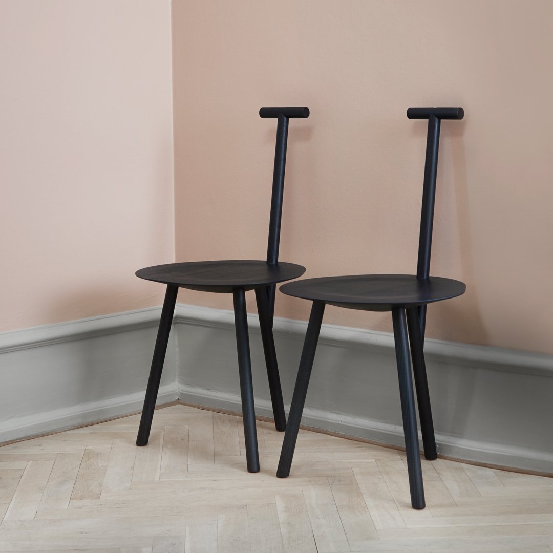 Black minimalist chairs with unique T-shaped backrests, set against a soft pink wall and herringbone floor pattern.