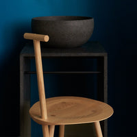 Natural wood chair with smooth curves, paired with a textured black bowl against a deep blue wall.