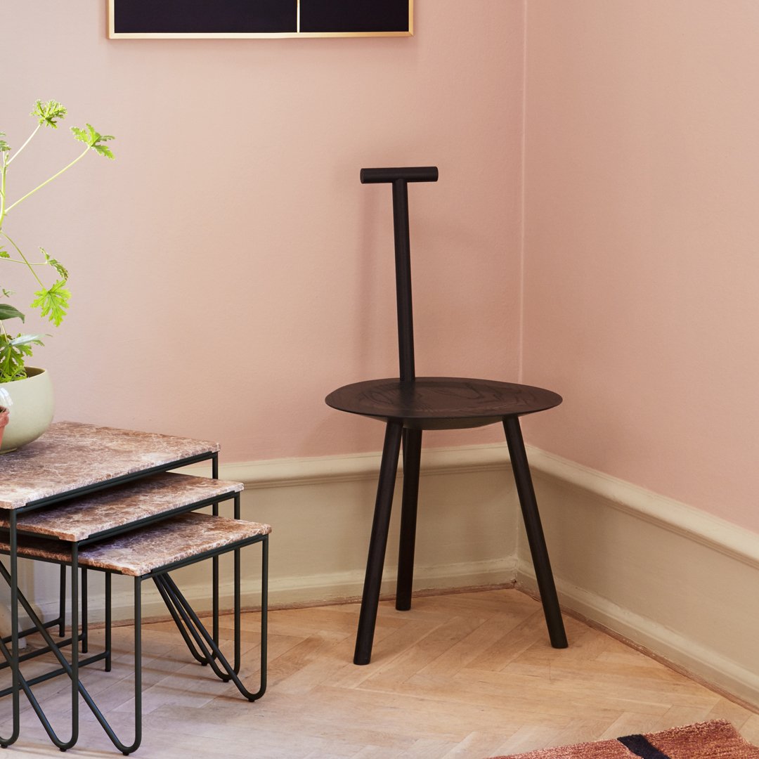 Black round stool with minimalist design, set against a soft pink wall and light wooden floor.