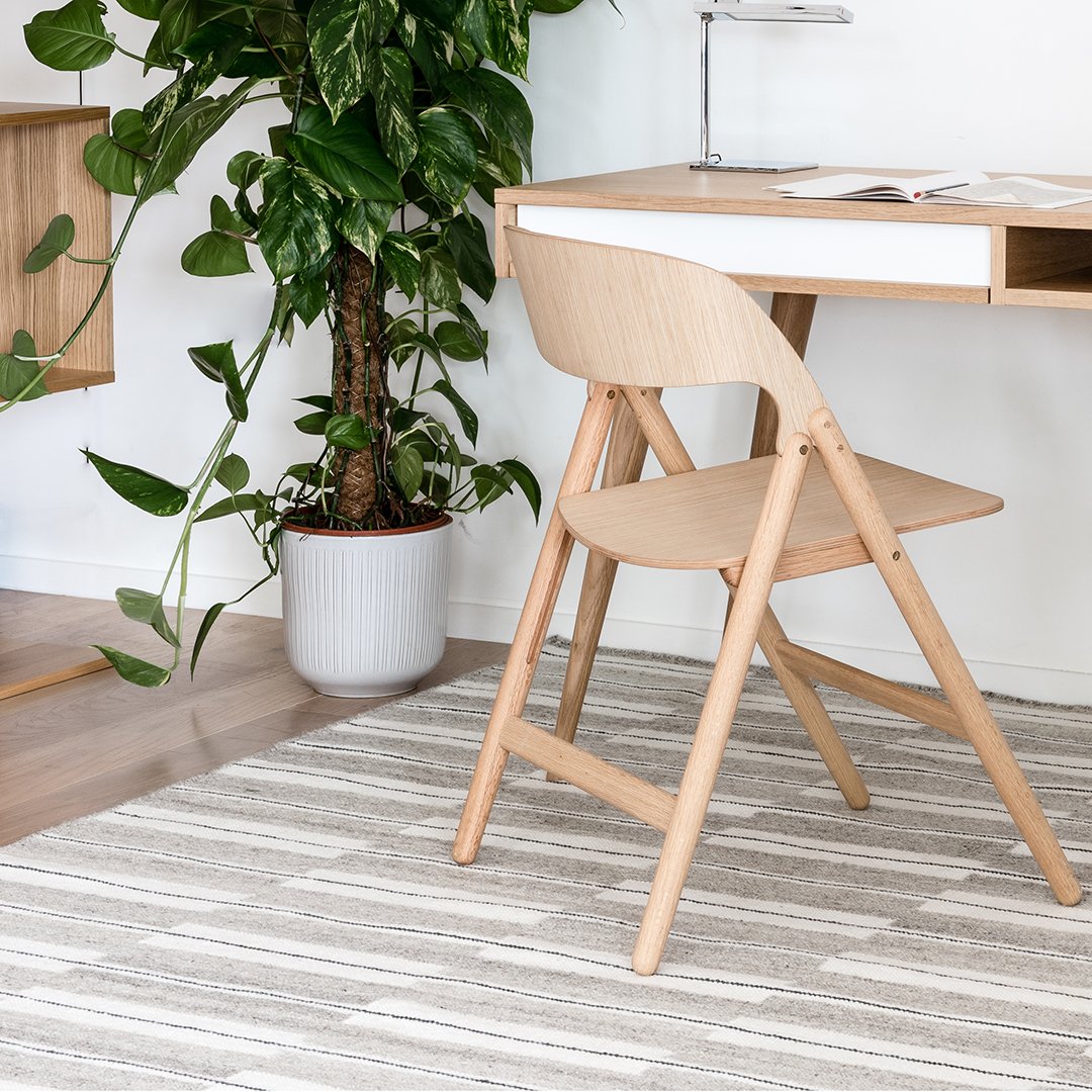 Light wood folding chair on a textured gray and white striped rug, with greenery in the background.