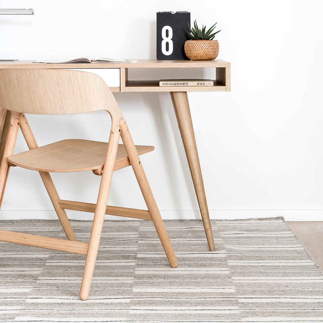 Light wood desk with angled legs, paired with a beige chair and a striped gray area rug, creating a minimalist aesthetic.