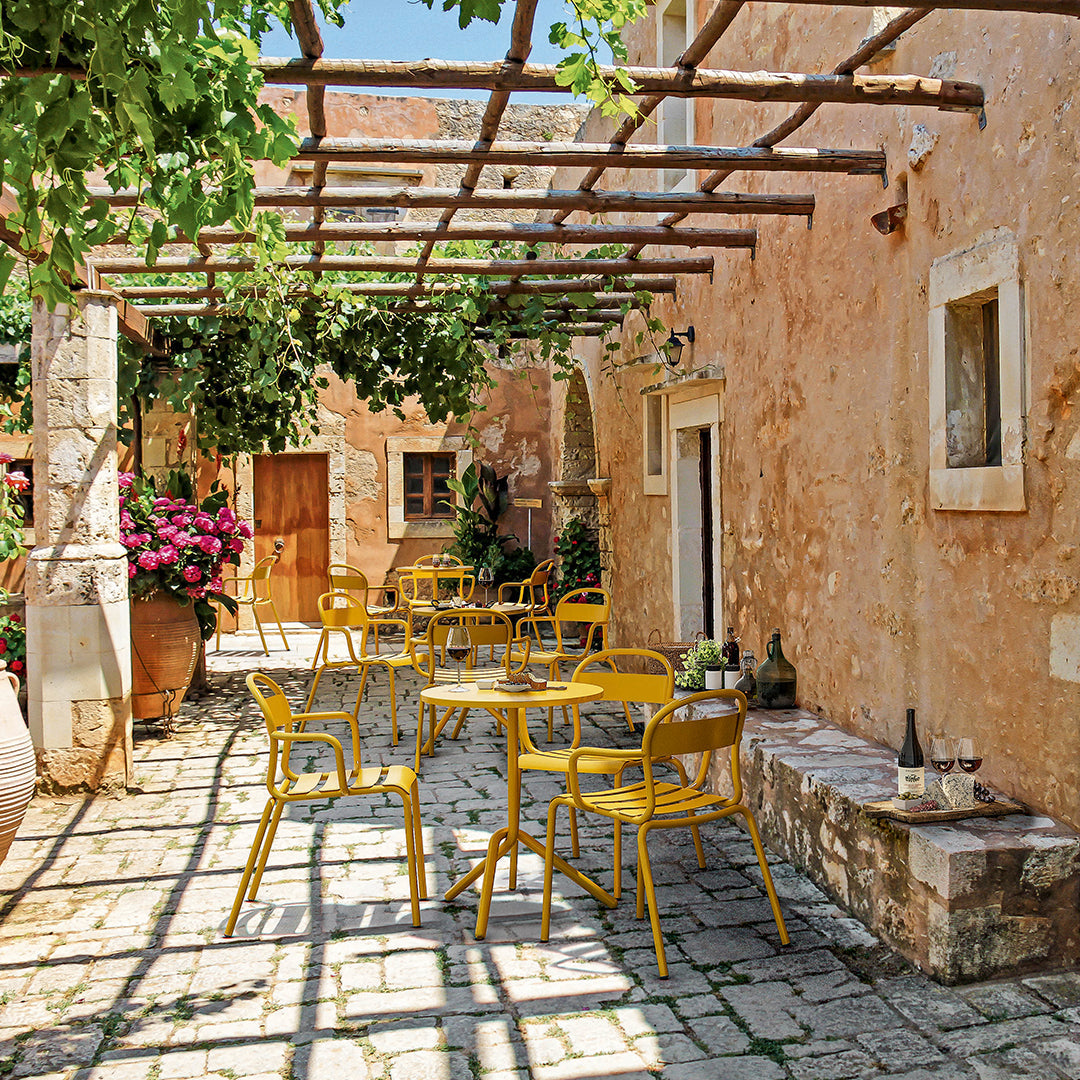 Bright yellow chairs against rustic stone walls, with green vines and vibrant flowers creating a lively outdoor setting.