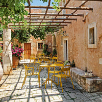 Bright yellow chairs against rustic stone walls, with green vines and vibrant flowers creating a lively outdoor setting.