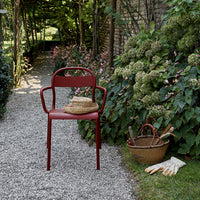 Red metal chair surrounded by lush green foliage and blooming flowers, set on a gravel pathway.