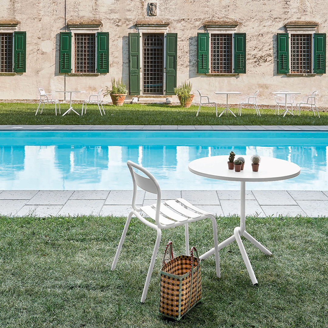 White round table and chair with a natural wicker basket, surrounded by green grass and a serene blue pool.