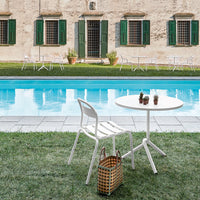 White round table and chair with a natural wicker basket, surrounded by green grass and a serene blue pool.