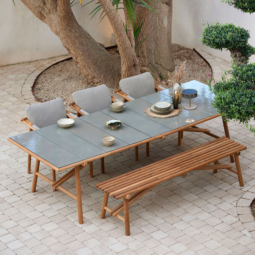 "Natural wood dining table with gray cushions, set against a sandy stone patio and lush greenery."