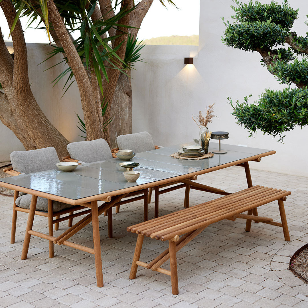 "Light wood dining table with gray cushions and a glass top, set against a natural stone patio and greenery."