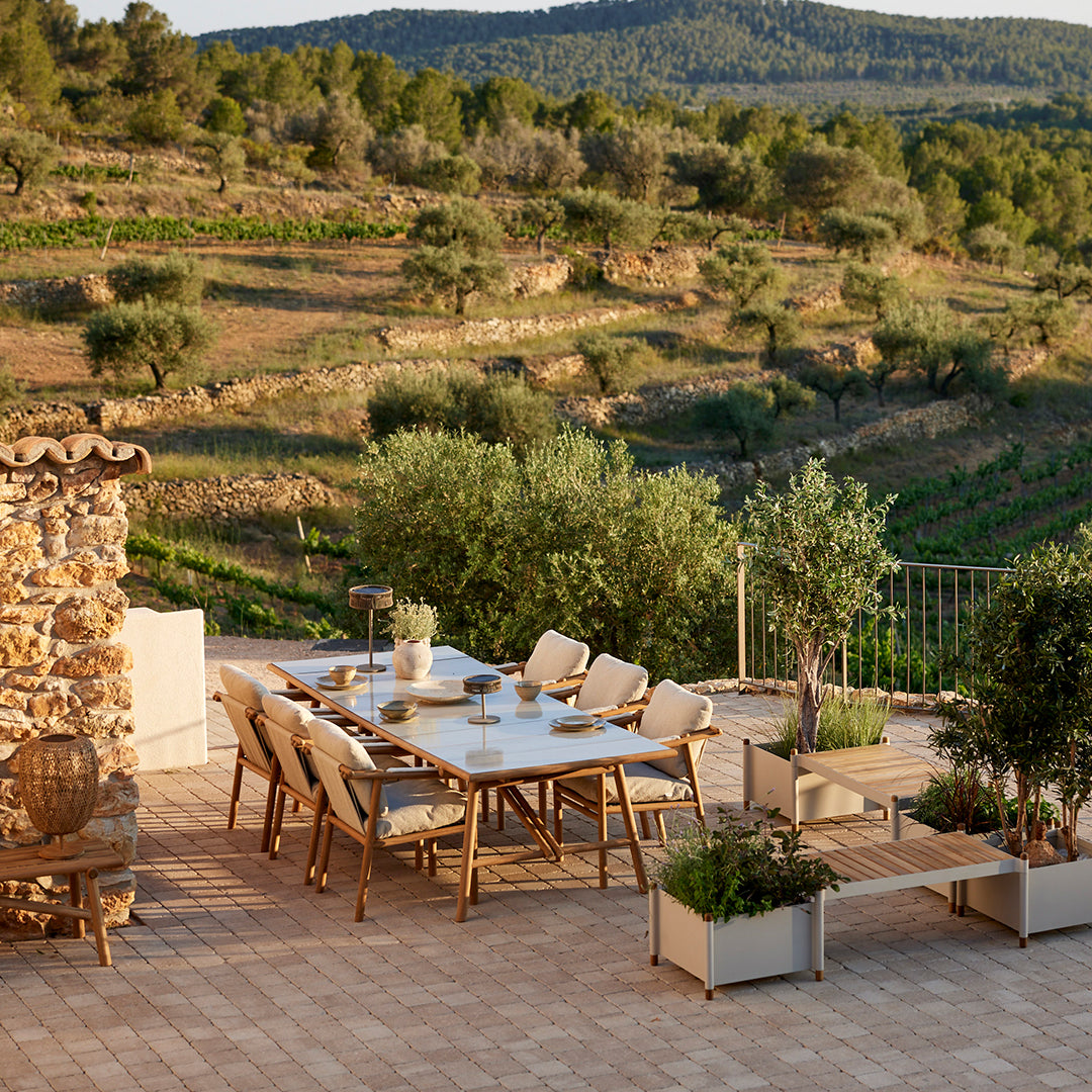 Outdoor dining set featuring beige cushions, natural wood tones, and green foliage against a rustic stone backdrop.