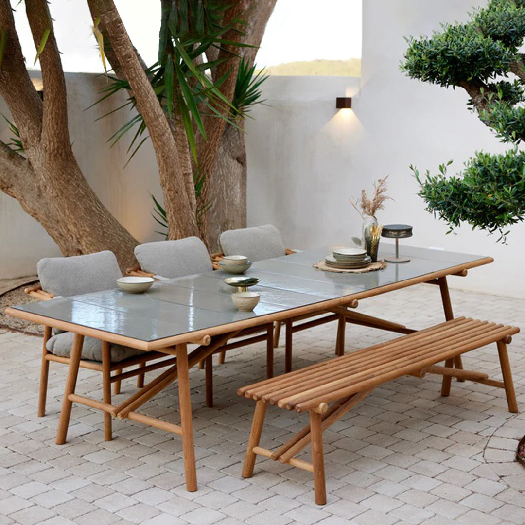Outdoor dining table set with gray tabletop, natural wood frame, and light gray cushions, surrounded by greenery.