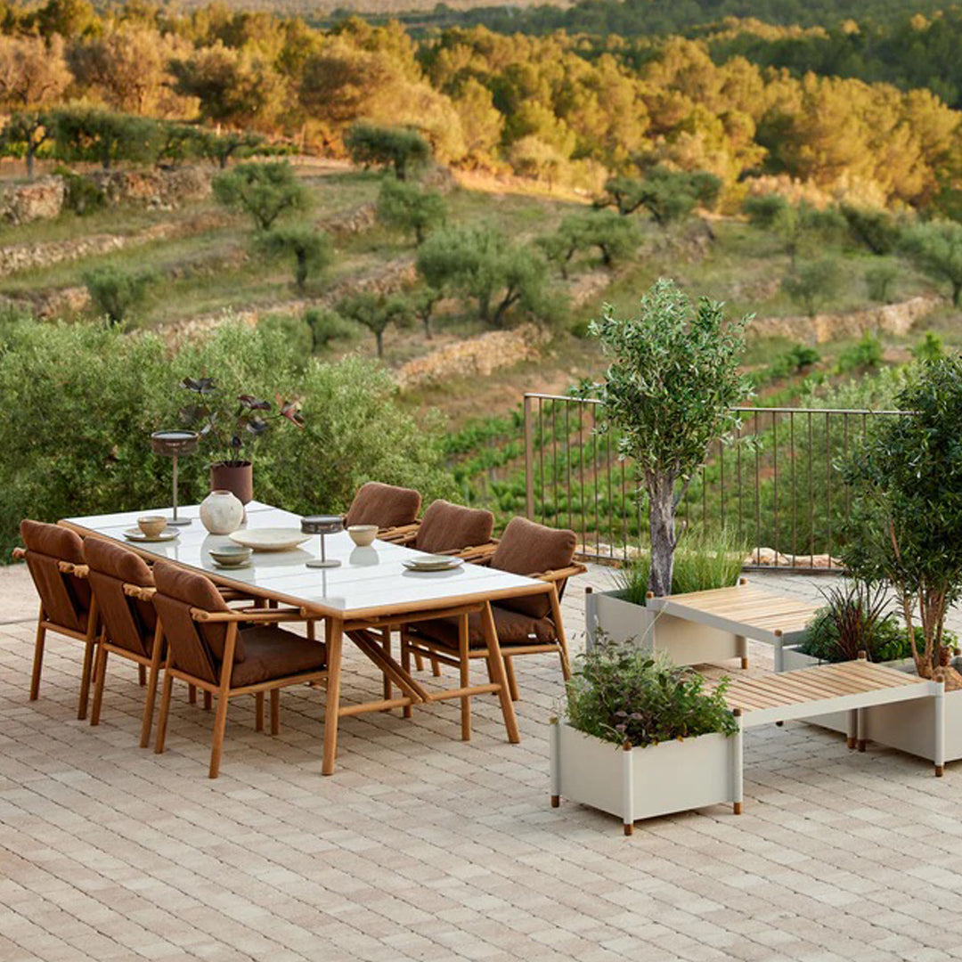 Brown outdoor dining set with a white table, green plants, and textured stone patio against a natural landscape backdrop.