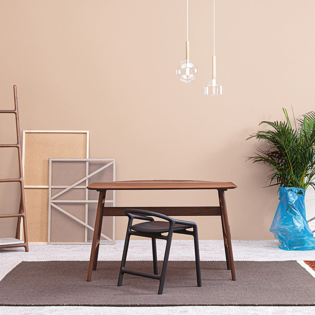Modern brown wood table with black chair, gray rug, and light beige wall; minimalistic design with green plant accents.
