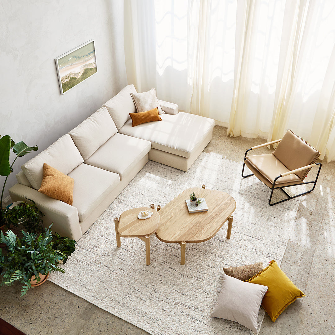 Beige sofa with orange and textured pillows, light wood furniture, and a cream rug in a bright, airy living room.