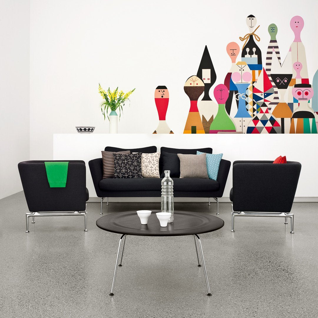 "Modern living room with black furniture, colorful patterned cushions, and a vibrant wall mural featuring abstract figures."