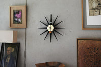 Black and gold starburst wall clock on a textured gray background with framed artwork. Retro design meets modern decor.