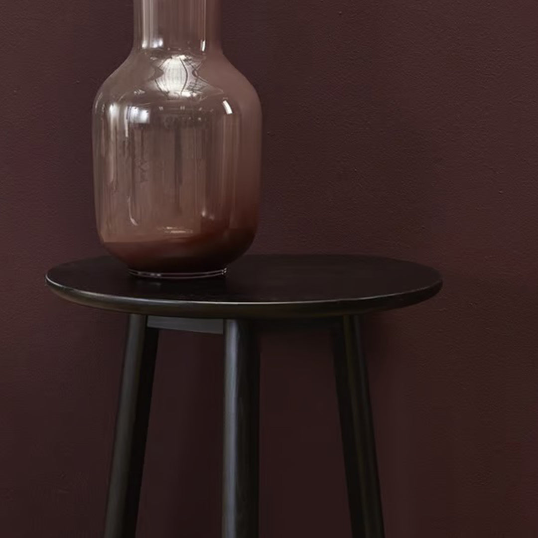 Black round side table with sleek legs, paired with a smooth, amber glass vase against a deep purple wall.