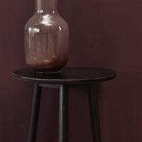Black round side table with sleek legs, paired with a smooth, amber glass vase against a deep purple wall.