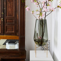 Large grey-green glass vase with pink cherry blossoms, set on a white pedestal beside a wood-paneled wall.